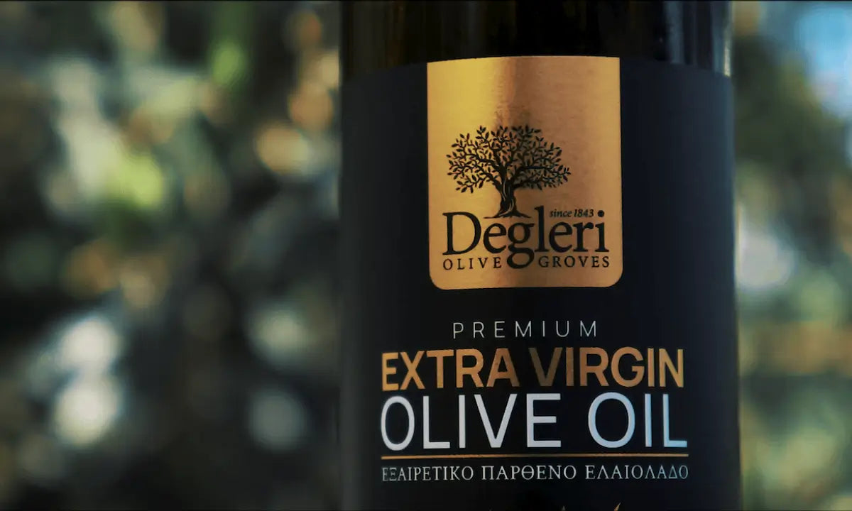 Close-up of a Degleri Premium Extra Virgin Olive Oil bottle label with the Degleri Olive Groves logo, set against a softly blurred background of olive trees and sunlight.