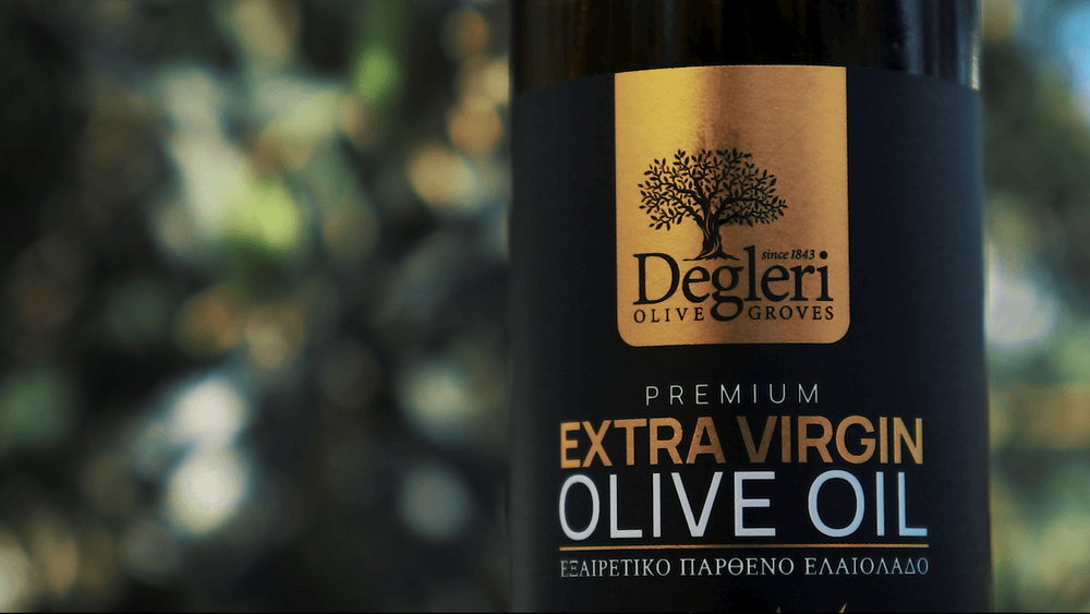 Close-up of a Degleri Premium Extra Virgin Olive Oil bottle label with the Degleri Olive Groves logo, set against a softly blurred background of olive trees and sunlight.