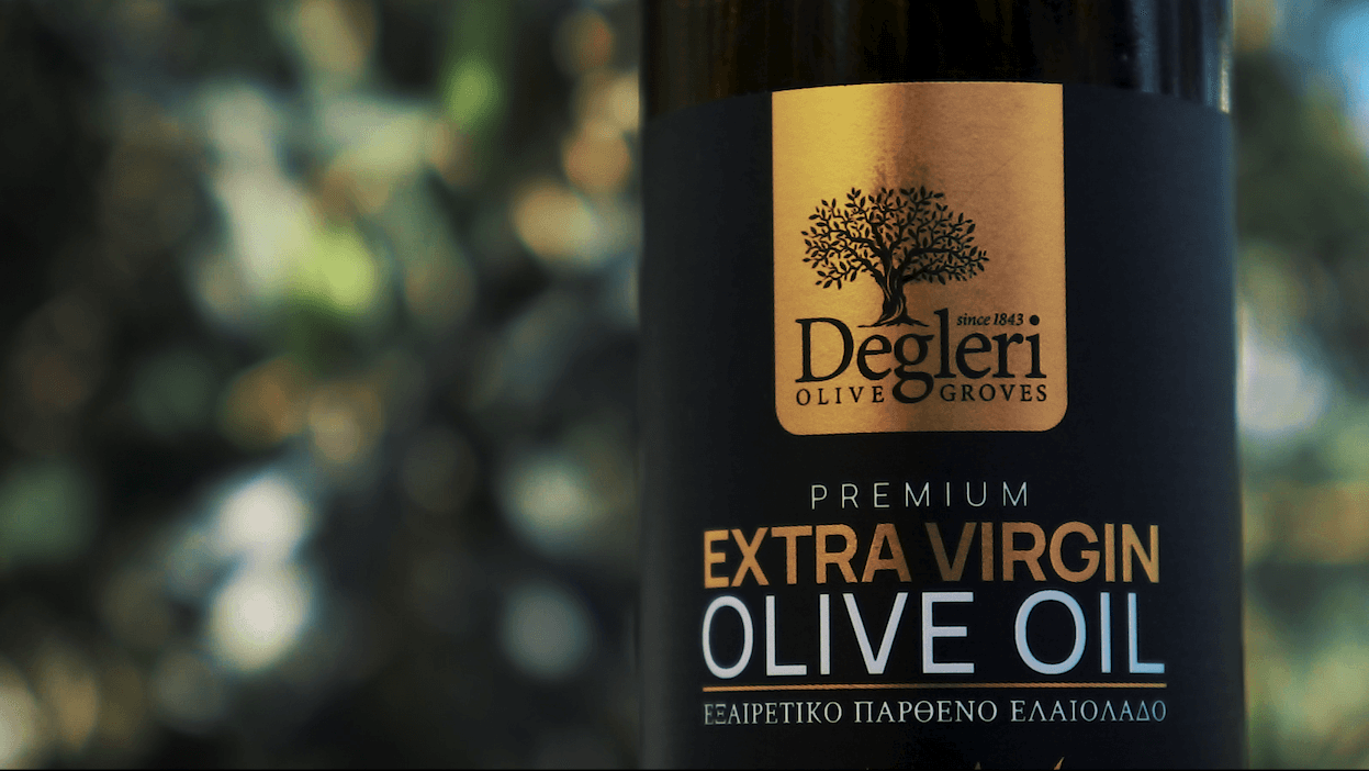 Close-up of a Degleri Premium Extra Virgin Olive Oil bottle label with the Degleri Olive Groves logo, set against a softly blurred background of olive trees and sunlight.